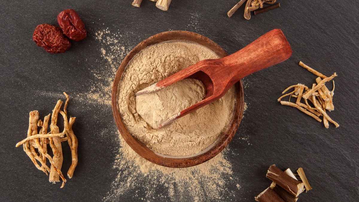 Mletá ashwagandha por v drevenej miske s lyžicou a sušenými koreňmi okolo.