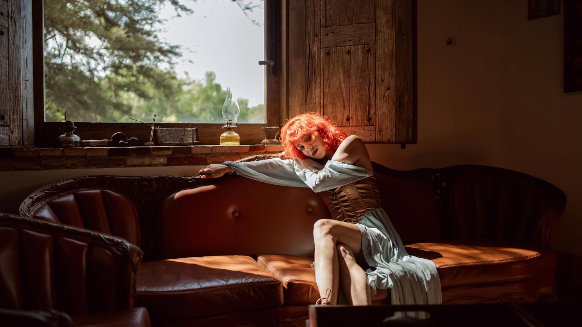 Žena s červenými vlasmi relaxuje na pohovke pri okne, svetlo preniká do miestnosti.