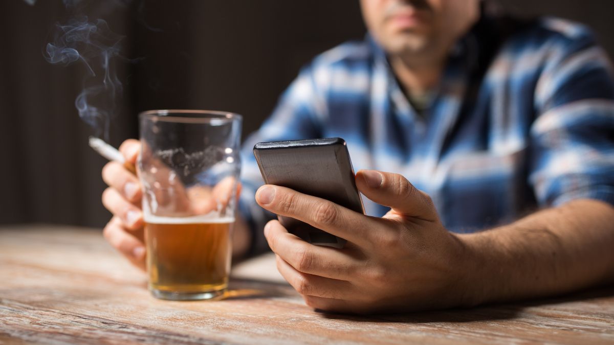 Muž drží pohár piva a mobilný telefón, dym z cigarety sa vznáša.